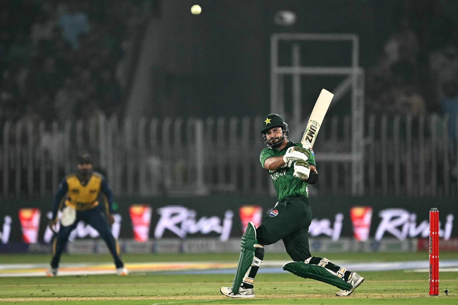 Sahibzada Farhan, a Pakistani cricketer in a green uniform, helmet, and pads, swings his bat at a cricket ball, which is suspended in mid-air. A wicketkeeper in a yellow and blue uniform is blurred in the background, and the stadium lights faintly illuminate the scene.