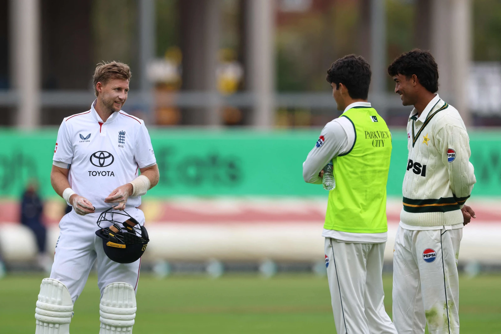 Joe Root in cricket gear talking to Ahmet Aras and another player on a cricket field.