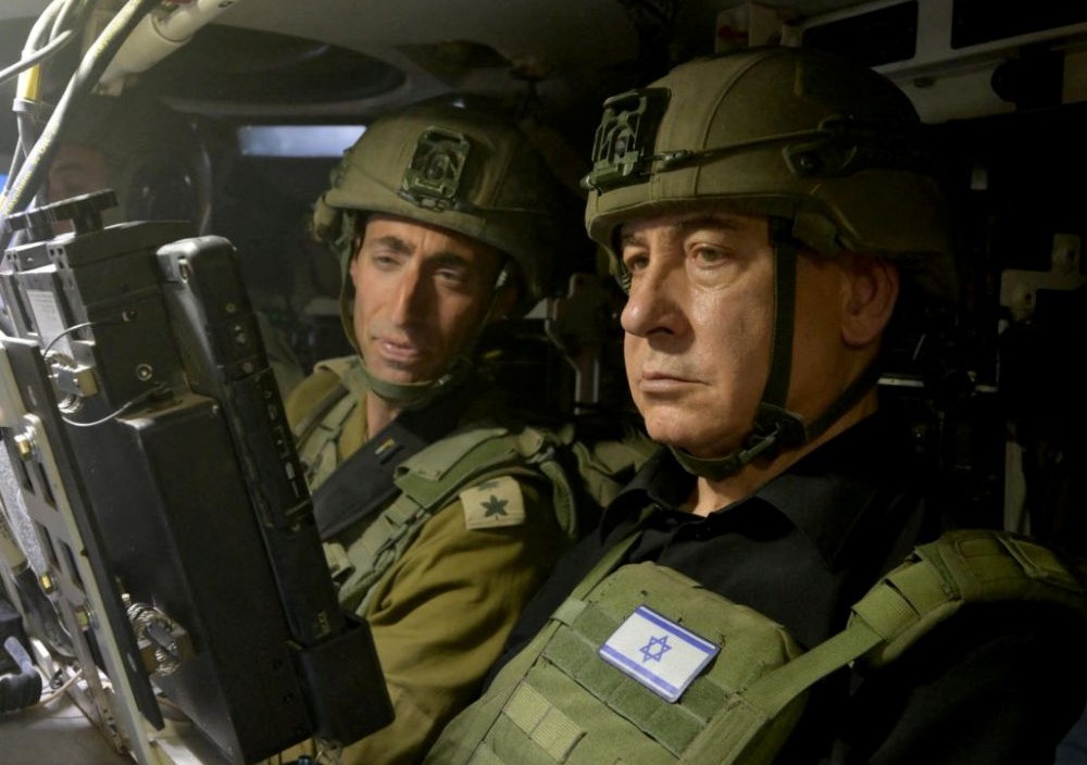 Two military personnel are inside a tank, wearing helmets and looking at a screen with focused expressions. They are in a confined, technology-filled space, and one is wearing a vest with an Israeli flag.