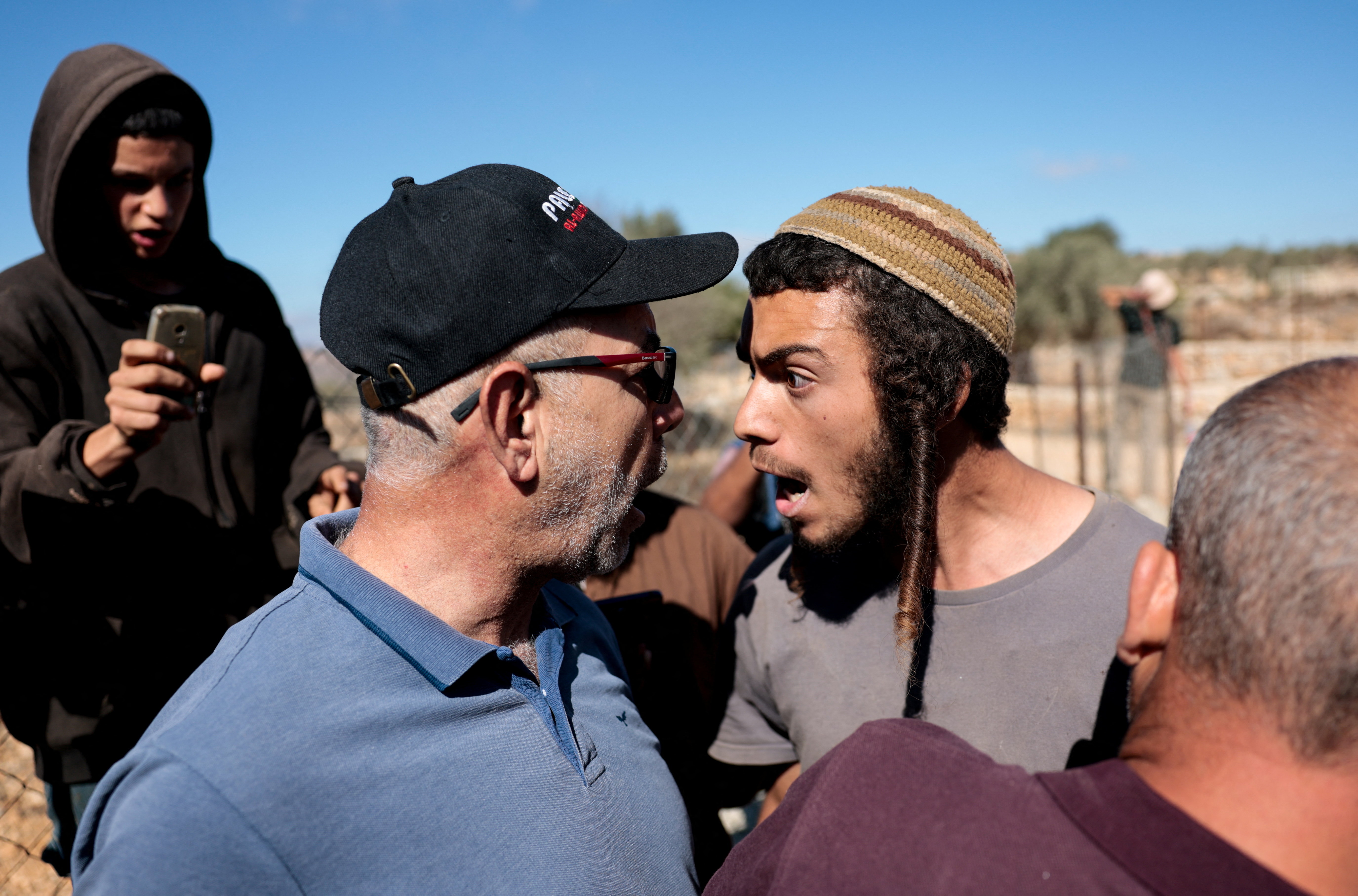 Tensions flare between two men during a heated dispute in a contested area, as onlookers capture the confrontation on their phones.