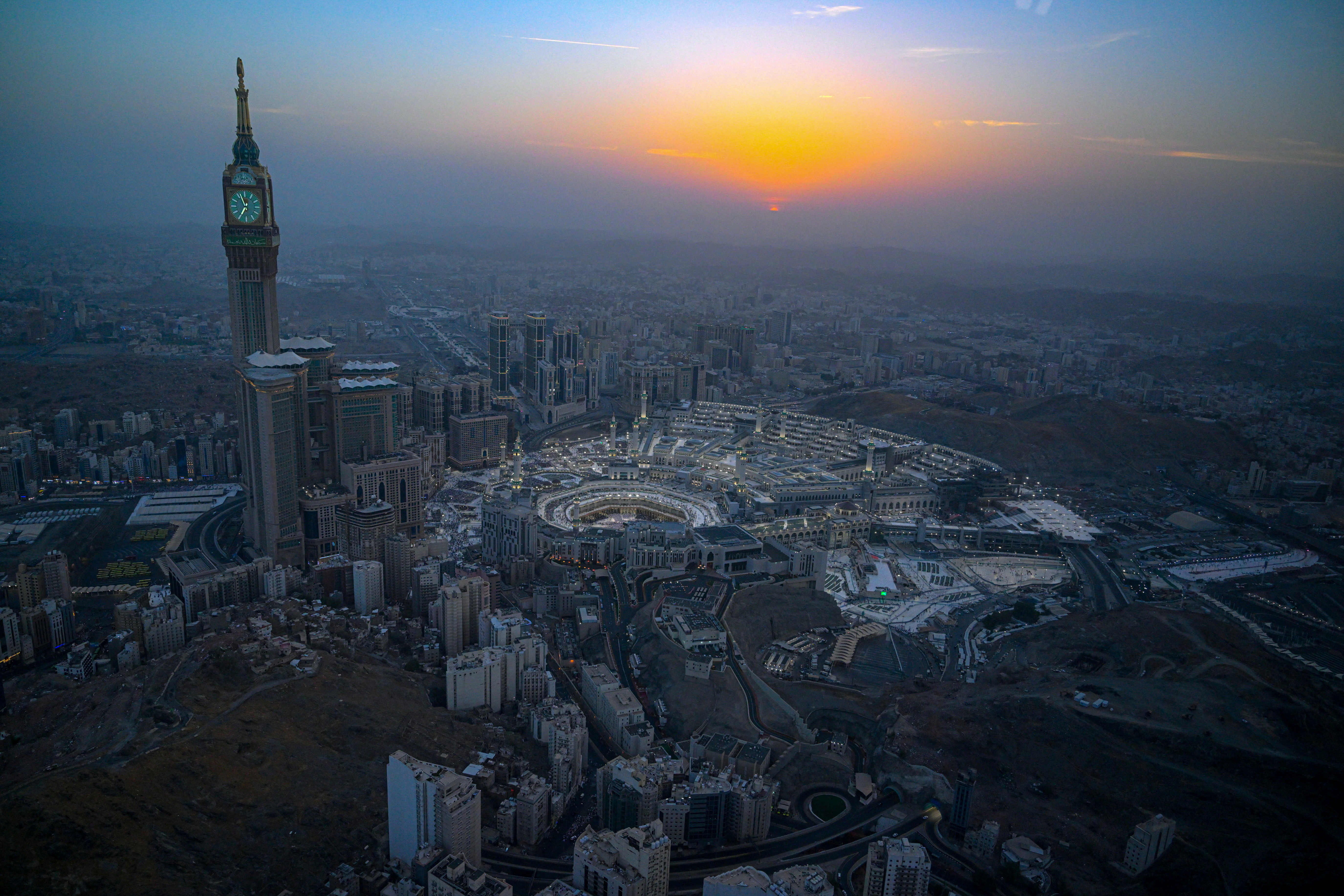 A stunning aerial view of a bustling cityscape at sunset, featuring a prominent clock tower and illuminated buildings surrounding a sacred site. The sky transitions from orange to deep blue, creating a serene and majestic atmosphere.
