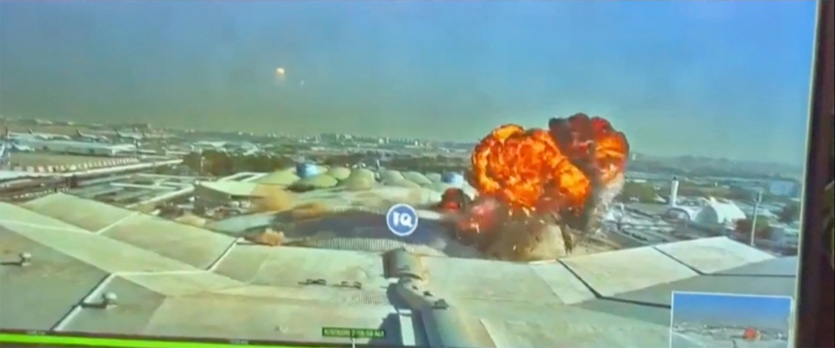 A large explosion erupts over a complex of buildings, possibly an airport or industrial facility, under a clear sky.