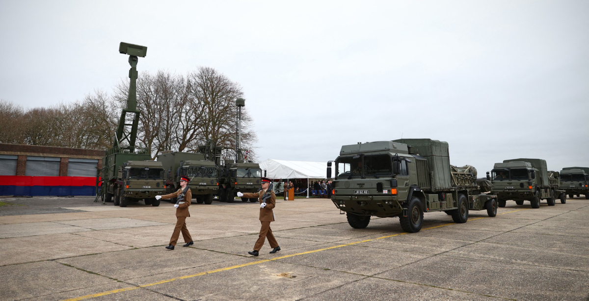 Soldiers march past a lineup of military trucks, some equipped with radar systems, during an outdoor event.