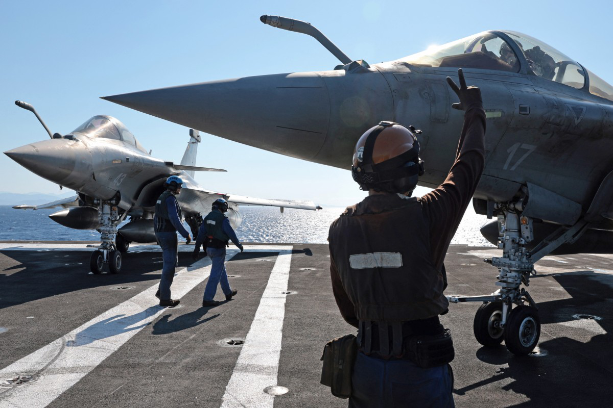 A flight deck officer signals to the pilot of a Dassault Rafale fighter jet as another aircraft stands by on an aircraft carrier at sea.