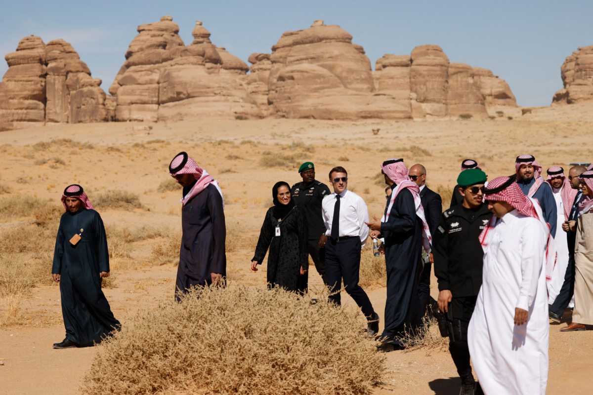 French President Emmanuel Macron walks with a group of individuals, many in traditional Saudi attire, through a desert landscape featuring large rock formations in the background.