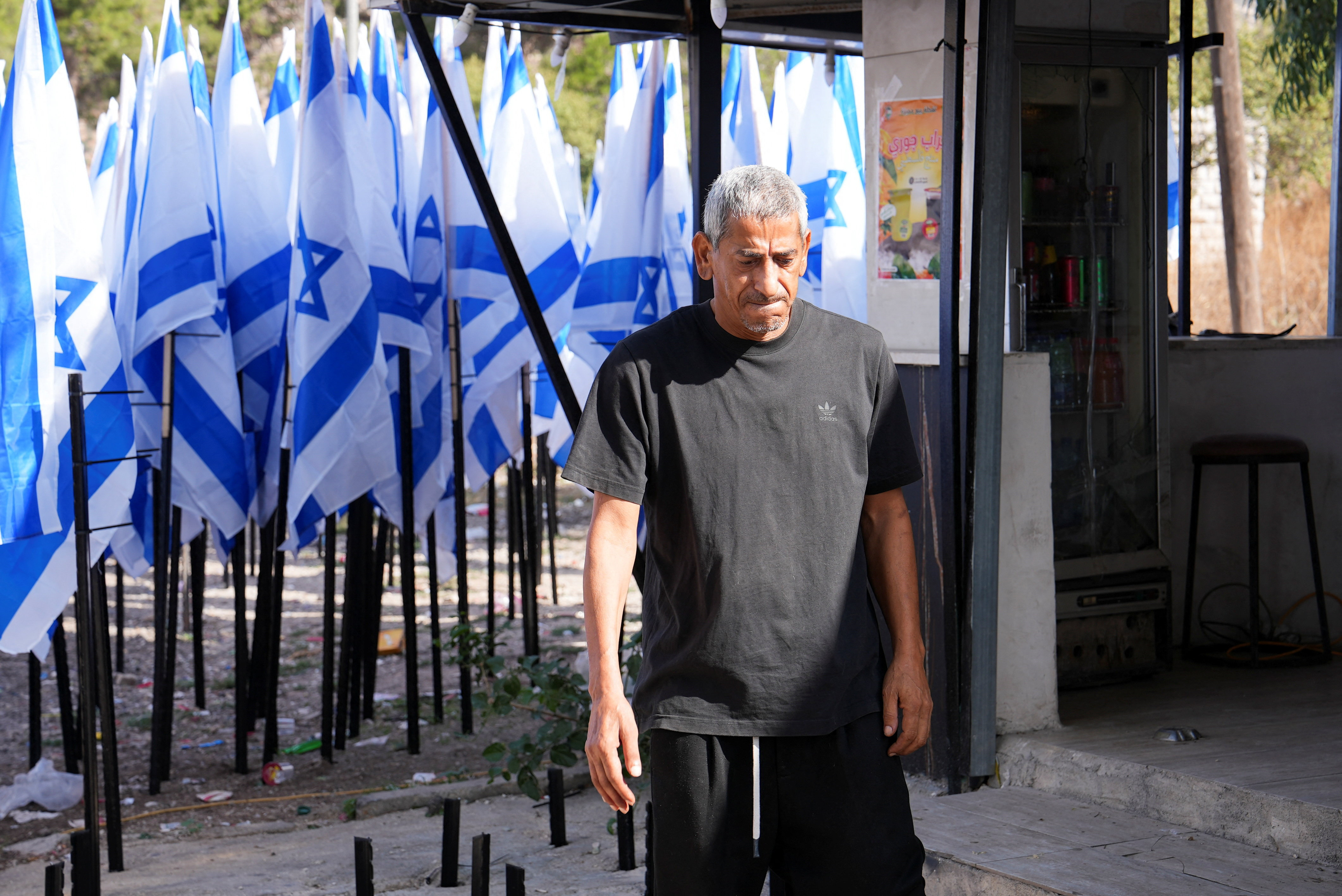 Palestinian coffee shop owner Mueyyid Hussein stands outside his business as Israeli flags erected by settlers stand nearby, which he says have obstructed the shop’s entrance and visibility. (Reuters)