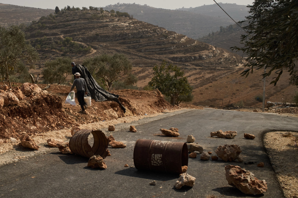 Palestinian men walk past a road, blocked in an effort to ward off Israeli landgrabbers, as they enter into an olive grove for harvest in the West Bank town of Sinjil, Oct. 24, 2026. (AP Photo/Leo Correa)