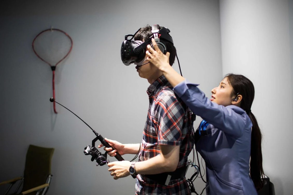 An operator adjusts the headset of a man as he plays a virtual reality (VR) fishing game at the Bandai Namco game facility of VR Zone Shinjuku in Tokyo on July 22, 2017. / AFP / Behrouz MEHRI