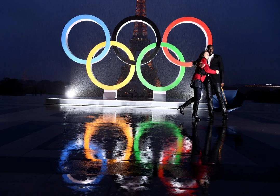A couple pose in the rain beside the Olympics Rings on the Trocadero Esplanade near the Eiffel Tower in Paris, on September 13, 2017, after the International Olympic Committee named Paris host city of the 2024 Summer Olympic Games. The International Olympic Committee named Paris and Los Angeles as hosts for the 2024 and 2028 Olympics on September 13, 2017, crowning two cities at the same time in a historic first for the embattled sports body. / AFP / CHRISTOPHE SIMON