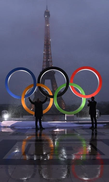 People gather beside The Olympics Rings as they are reflected in rainwater on the Trocadero Esplanade near the Eiffel Tower in Paris, on September 13, 2017, after the International Olympic Committee named Paris host city of the 2024 Summer Olympic Games. The International Olympic Committee named Paris and Los Angeles as hosts for the 2024 and 2028 Olympics on September 13, 2017, crowning two cities at the same time in a historic first for the embattled sports body. / AFP / CHRISTOPHE SIMON
