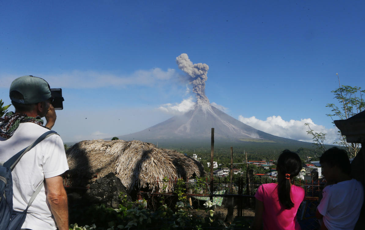 A foreign tourist, left, takes photos of an erupting Mayon volcano Tuesday, Jan. 23, 2018 as seen from Legazpi city, Albay province, southeast of Manila, Philippines. The Philippines' most active volcano ejected a huge column of lava fragments, ash and smoke in another thunderous explosion at dawn Tuesday, sending thousands of villagers back to evacuation centers and prompting a warning that a violent eruption may be imminent. (AP Photo/Bullit Marquez)