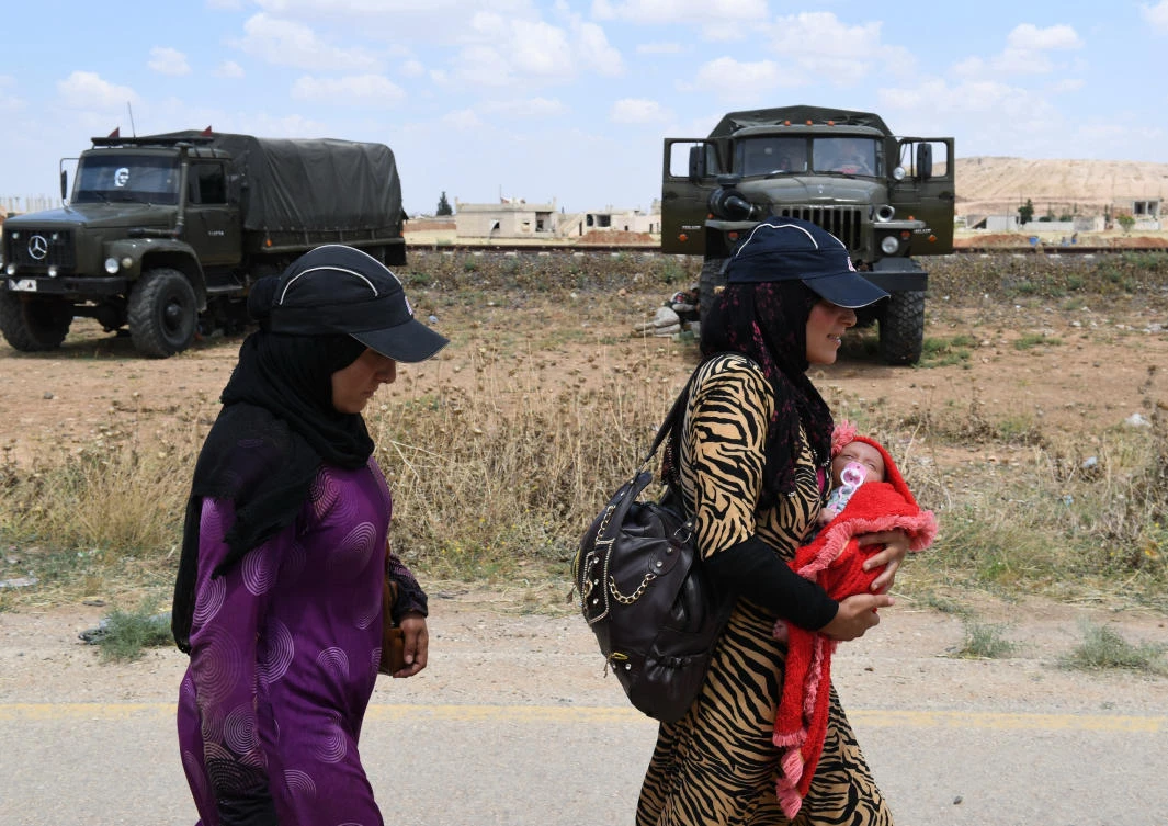 A Syrian woman walks carrying a child with another woman past military vehicles, after arriving in a convoy carrying displaced people into government-controlled territory at Abu al-Zuhur checkpoint in the western countryside of Idlib province, on June 1, 2018. (AFP)