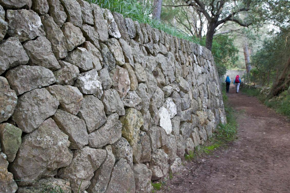 Two people walk past a wall built using the dry stone technique in Esporles on the Spanish Balearic Island of Mallorca on December 11, 2018. The art of dry stone walling, inscribed in 2018 on the Representative List of the Intangible Cultural Heritage of Humanity, (sometimes called drystack) is a building method by which structures are constructed from stones without any mortar to bind them together. Dry stone structures are stable because of their unique construction method, which is characterized by the presence of a load-bearing facade of carefully selected interlocking stones. / AFP / JAIME REINA