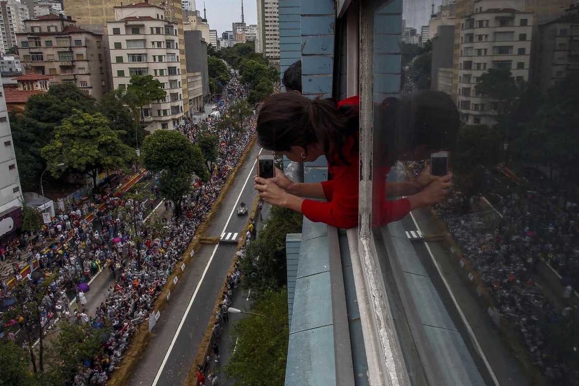 A woman leans out of a window to take pictures of the Red Bull Soapbox race in Sao Paulo, Brazil on April 14, 2019. The Red Bull Soapbox race is an annual event where amateur drivers race with their homemade soapbox vehicles down a 420 m hill through obstacles. / AFP / Miguel SCHINCARIOL