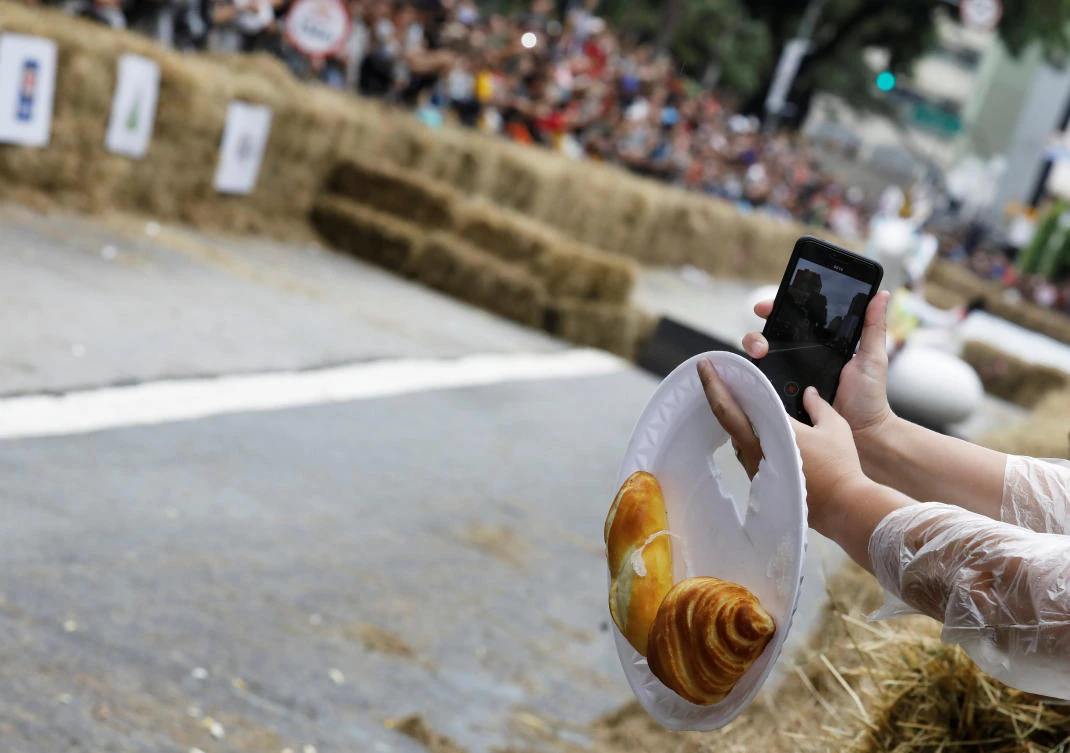 A man holds a plate with a plastic bread and croissant as he records competitors riding a home-made vehicle on a downhill track during the Red Bull Soapbox Race in Sao Paulo, Brazil, April 14, 2019. REUTERS/Nacho Doce