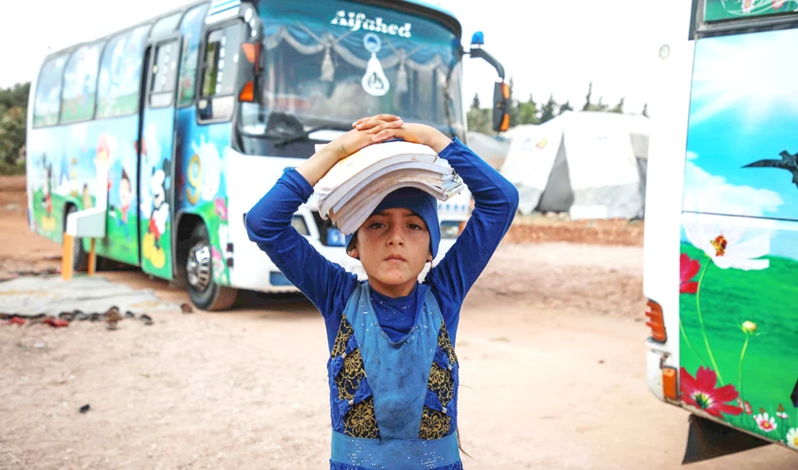 A displaced Syrian girl carries books on her head near a bus converted into a classroom in the village of Hazano in northwestern Syria. (AFP)
