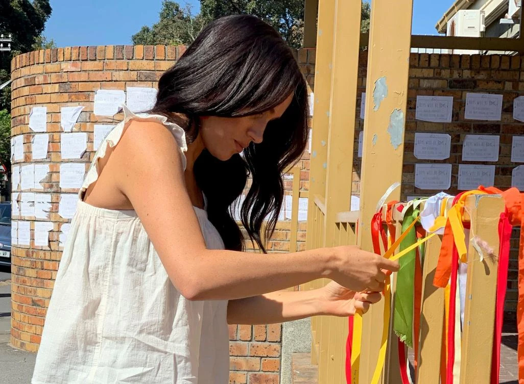 A recent undated handout photograph released on September 28, 2019 by the Duke and Duchess of Sussex shows Britain's Meghan, Duchess of Sussex ties on a ribbon as she visits the memorial to murdered South African student Uyinene Mrwetyana in Cape Town. (AFP)