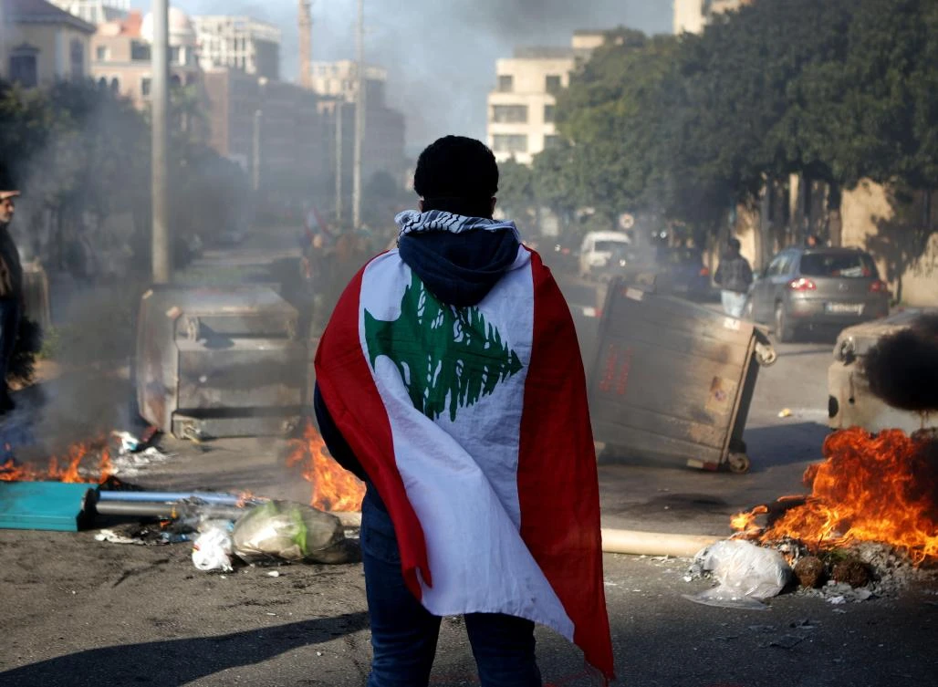A Lebanese anti-government protester, wrapped in a national flag, stands in front of a road blocked with burning tyres and overtunrned garbage dumpsteres in Beirut, on January 14, 2020. (AFP)
