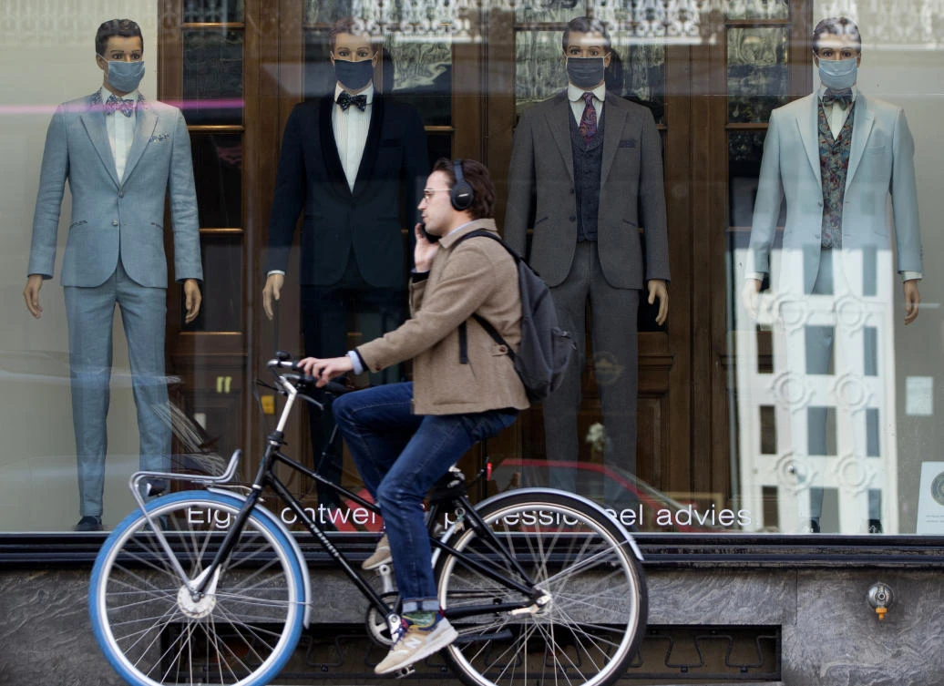 A man speaks on a mobile phone as he rides his bike past a men’s clothing store, with mannequins wearing mouth masks to protect against coronavirus that match their outfits, in Antwerp, Friday, March 27, 2020. (AP/Virginia Mayo)
