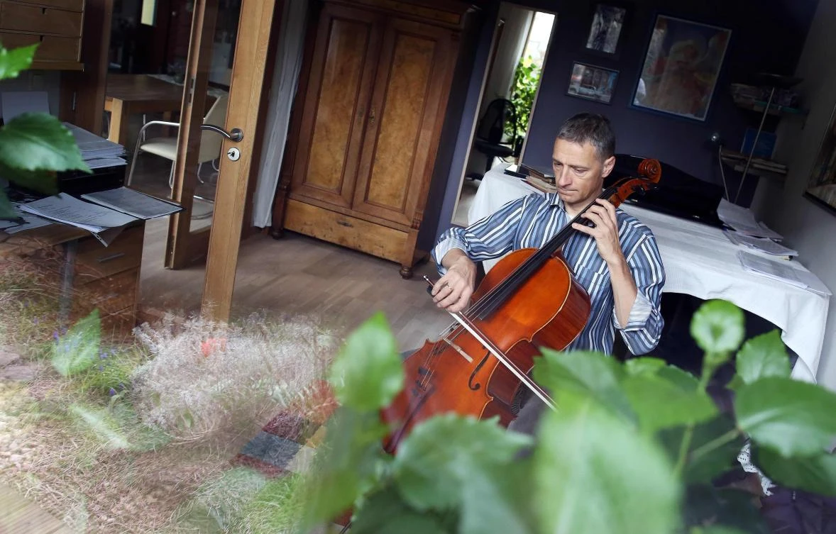 German musician Stephan Wuensch, principal cellist of Leipzig's opera house (Oper Leipzig), is seen through a window as he plays the violoncello at home, where he stays confined with his family due to the pandemic of the novel coronavirus, on April 3, 2020 on the outskirts of Leipzig, eastern Germany. The gates of the Leipzig Opera have been closed for about three weeks due to the novel corona pandemic - but the voices and instruments of the artists are far from silent. / AFP / Ronny Hartmann