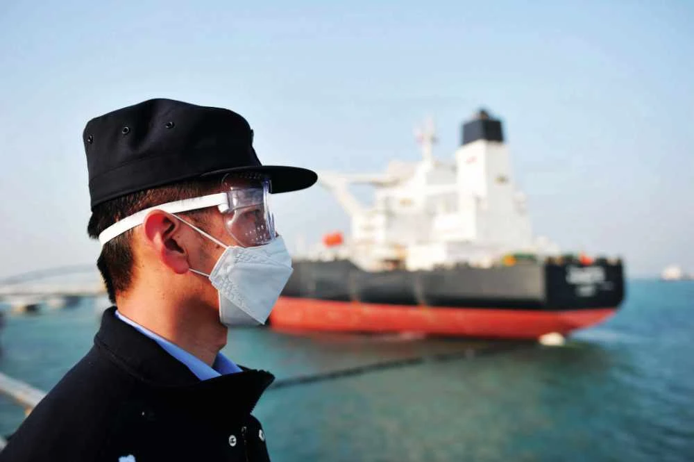 A police officer wearing a protective mask keeps watch as a Kuwaiti oil tanker unloads crude oil at the port of Qingdao, in China. Crude prices have fallen by up to two-thirds following the introduction of lockdowns to curb the spread of coronavirus. (AFP)