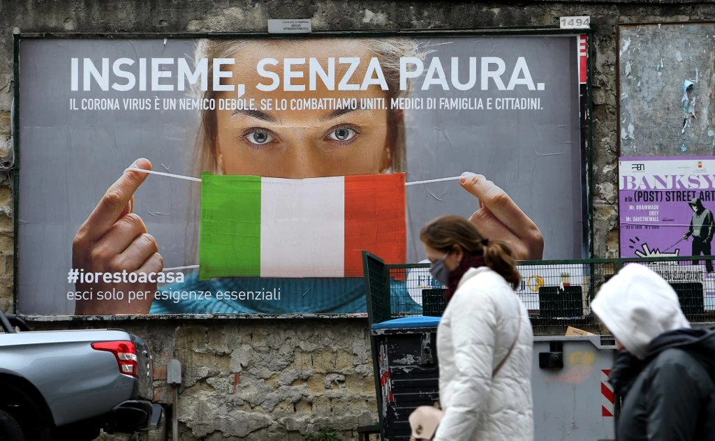 People walk past a huge billboard that shows a woman with as protective mask in the colours of an Italian flag and which reads, "All together, without fear" referring to the coronavirus campain to stay home in the city of Naples. (File/AFP)