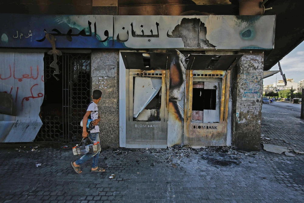 A youth walks with a shoeshine kit past a burnt down branch of a Lebanese bank after it was set on fire and vandalised by protesters earlier, in Al-Nour Square in Lebanon's northern port city of Tripoli on June 12, 2020. (AFP/File Photo)