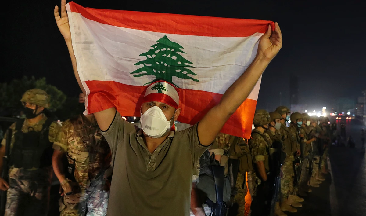 An anti-government protester holds up a Lebanese flag as army soldiers stand guard during a demonstration against deteriorating economic conditions as politicians are deadlocked over forming a new government, in the town of Jal El-Dib, north of Beirut, Lebanon, Sunday, Sept. 27, 2020. (AP)