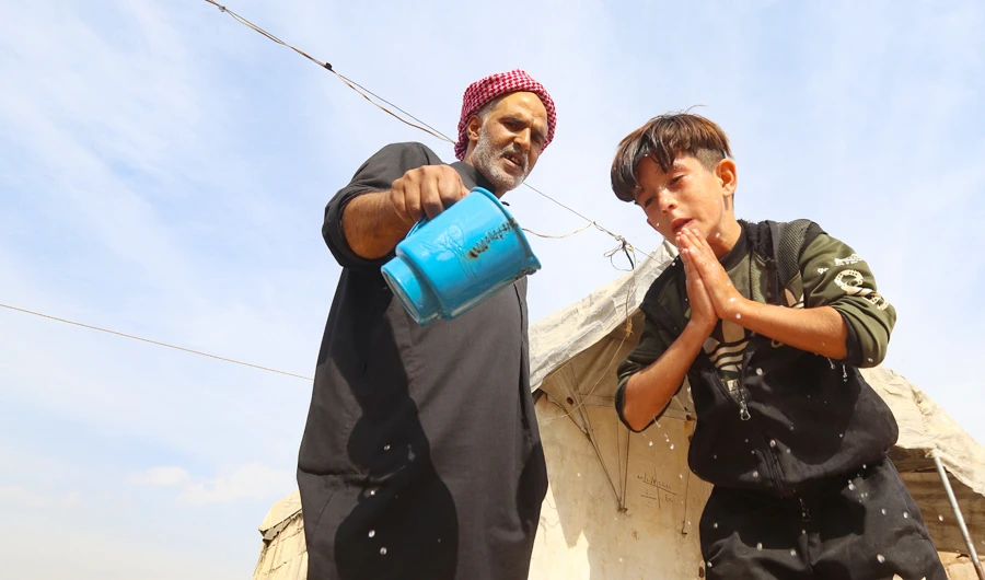 An elderly man helps a young boy wash his face in an overcrowded displacement camp near the village of Qah in Syria’s northwestern Idlib province, during the novel coronavirus pandemic crisis. (AFP)