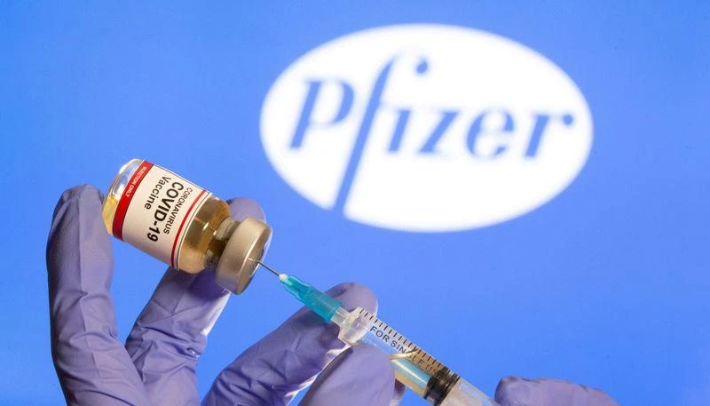A woman holds a small bottle labeled with a “Coronavirus COVID-19 Vaccine” sticker and a medical syringe in front of displayed Pfizer logo in this illustration taken. (File/Reuters)