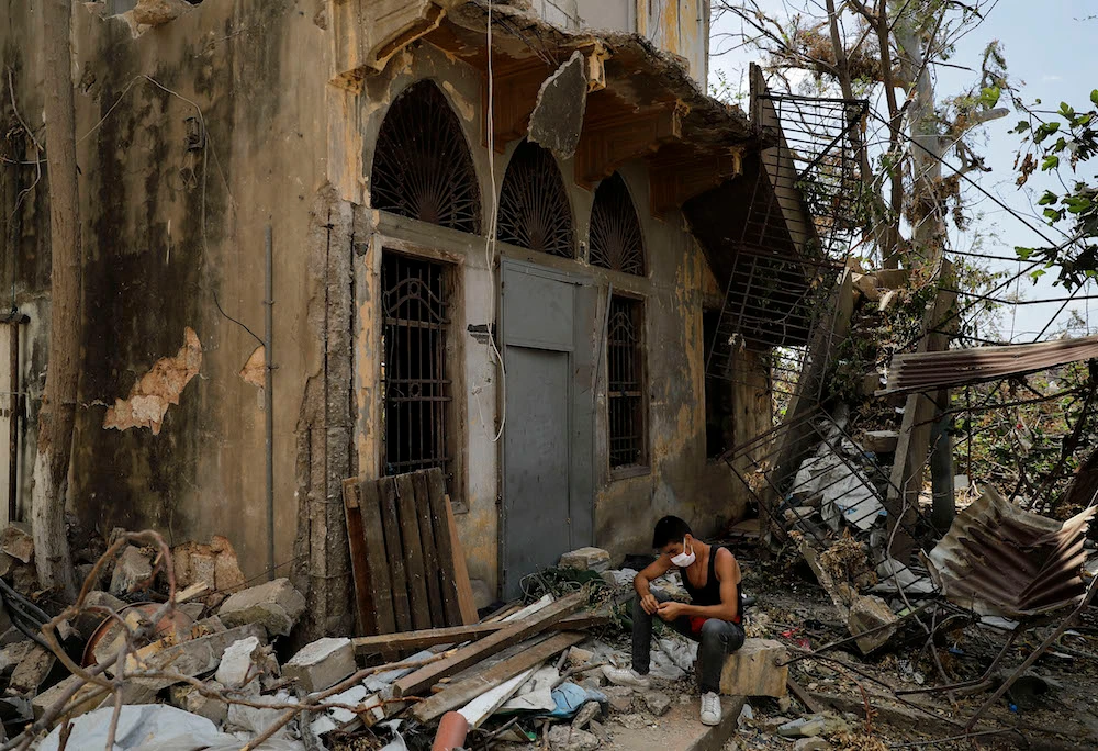 A Syrian national sits next to heavily damaged buildings in Beirut’s Karantina neighborhood almost two weeks after a massive explosion at the city’s port shook the Lebanese capital. Six months after a massive explosion ripped through Beirut, donors say that most of the emergency aid they pledged for Lebanon’s needy has been disbursed. (File/AFP)