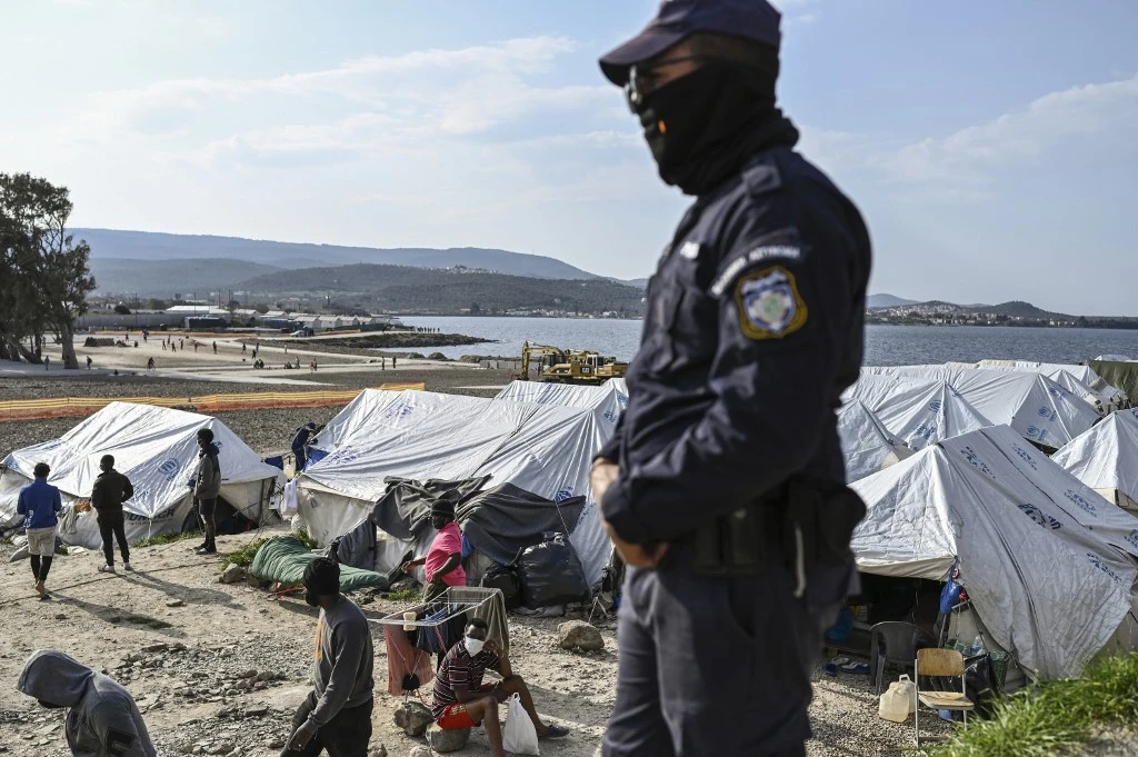 A Greek policeman stands in foreground as migrants walk inside the new refugee camp of Kara Tepe in Mytilene, on Lesbos, on March 29, 2021. (File/AFP)