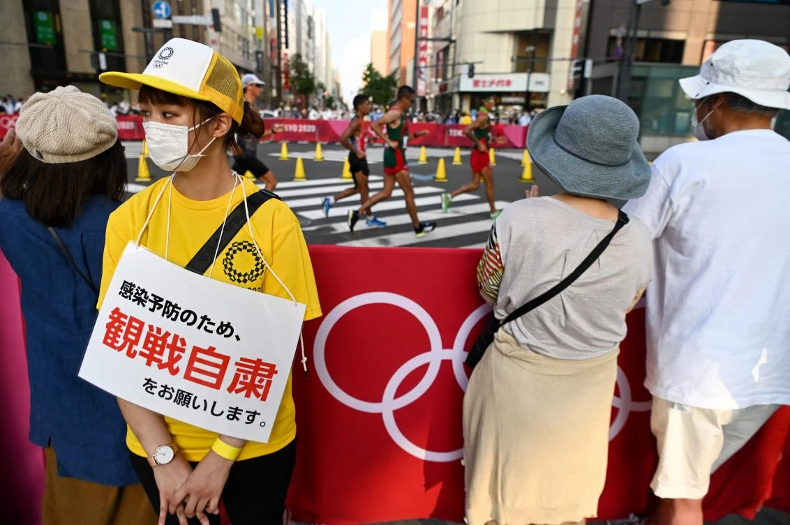 A volunteer wearing a sign reading “Don’t stop to watch the race due to the coronavirus disease restrictions” stands by people gathered to watch the men’s 20km race walk final during the Tokyo 2020 Olympic Games on Aug. 5, 2021. (AFP)