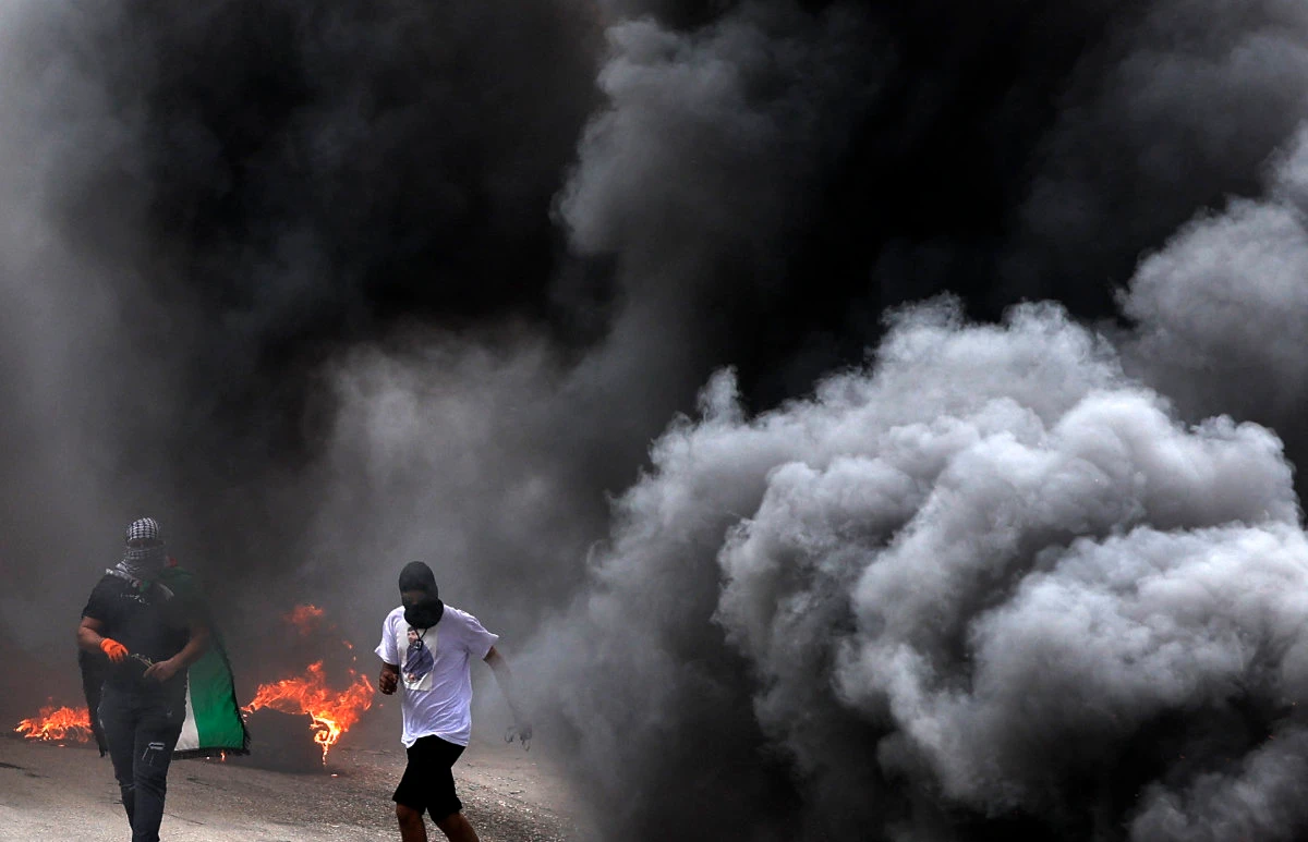 Palestinian protesters take cover behind smoke during clashes with Israeli forces in the village of Burqa, in the occupied West Bank, on April 19, 2022. (JAAFAR ASHTIYEH / AFP)