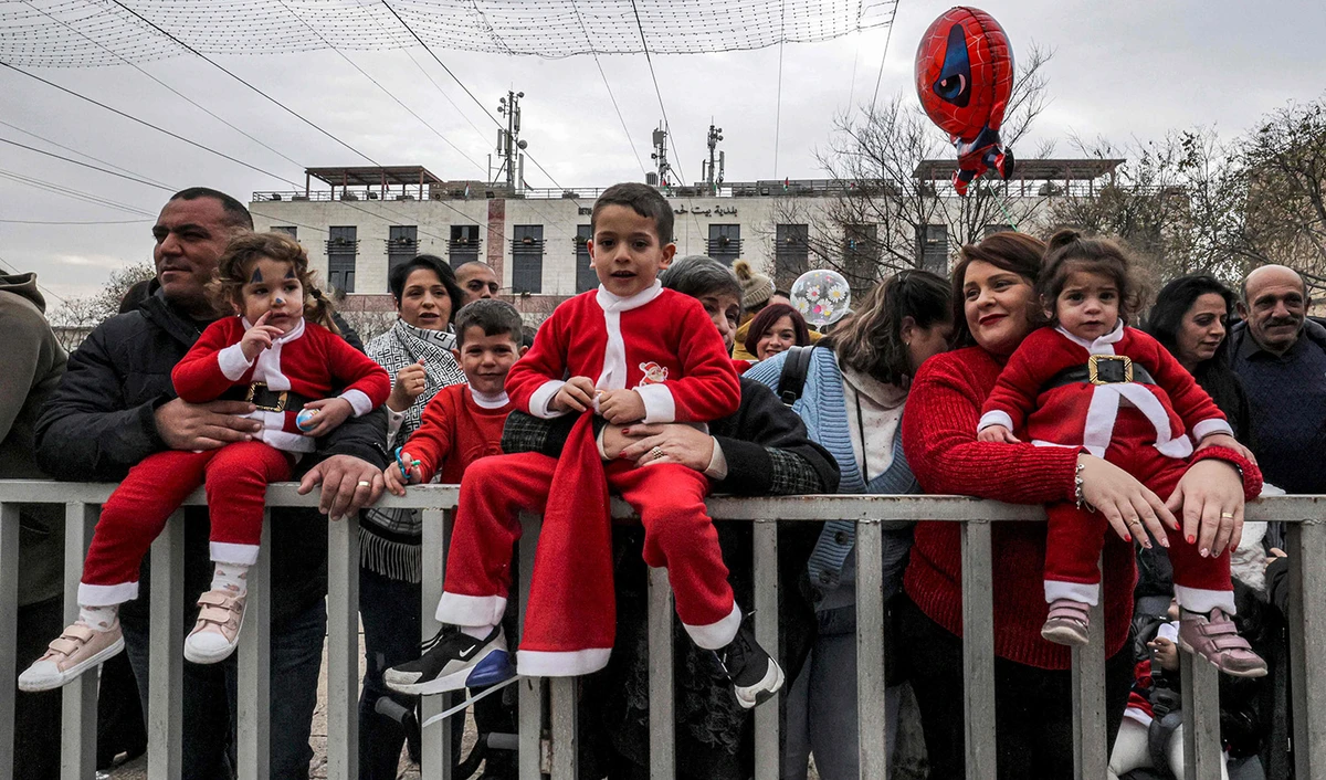 Children dressed in Christmas-themed clothing are held by adults as they sit on a fence to watch Christmas eve celebrations at the Manger Square outside the Church of the Nativity in the biblical city of Bethlehem in the occupied West Bank on December 24, 2022. (AFP)
