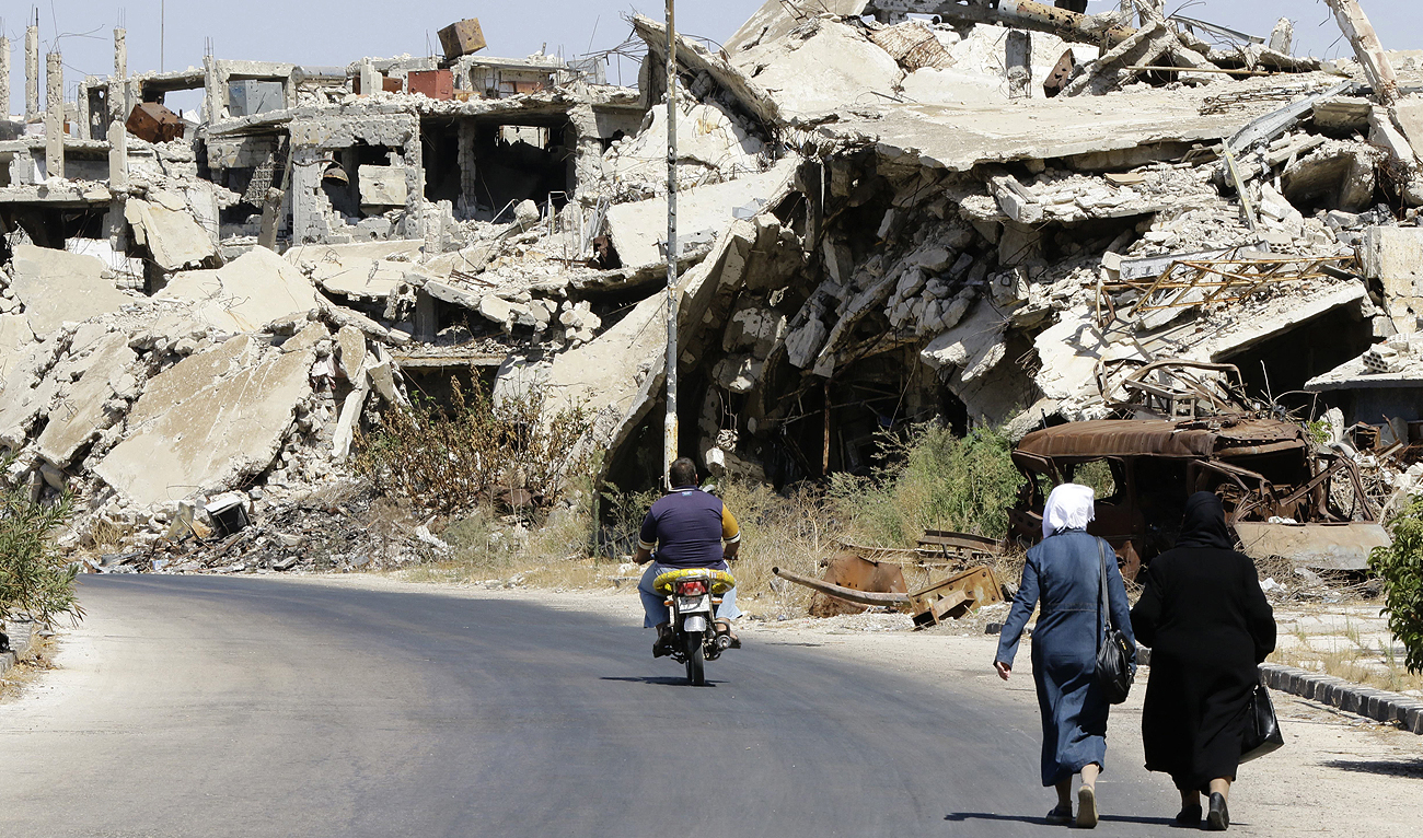 In this file picture, taken on September 19, 2016, Syrians walk and drive past destroyed buildings in Jouret al-Shiah neighbourhood of the central Syrian city of Homs. (Photo courtesy: AFP/File)
