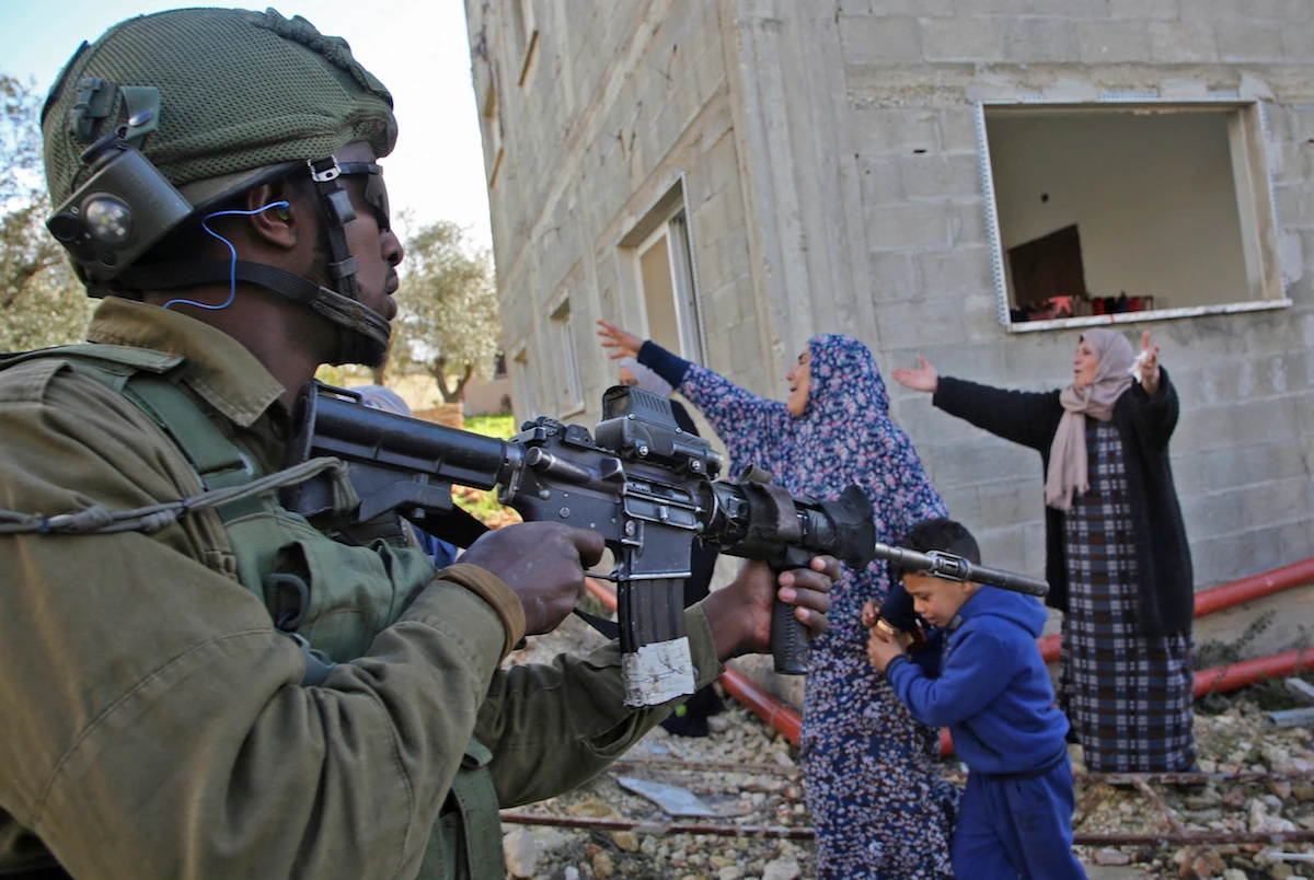 An Israeli soldier stands guard holding an assault rifle as Palestinian women protest. (File/AFP)