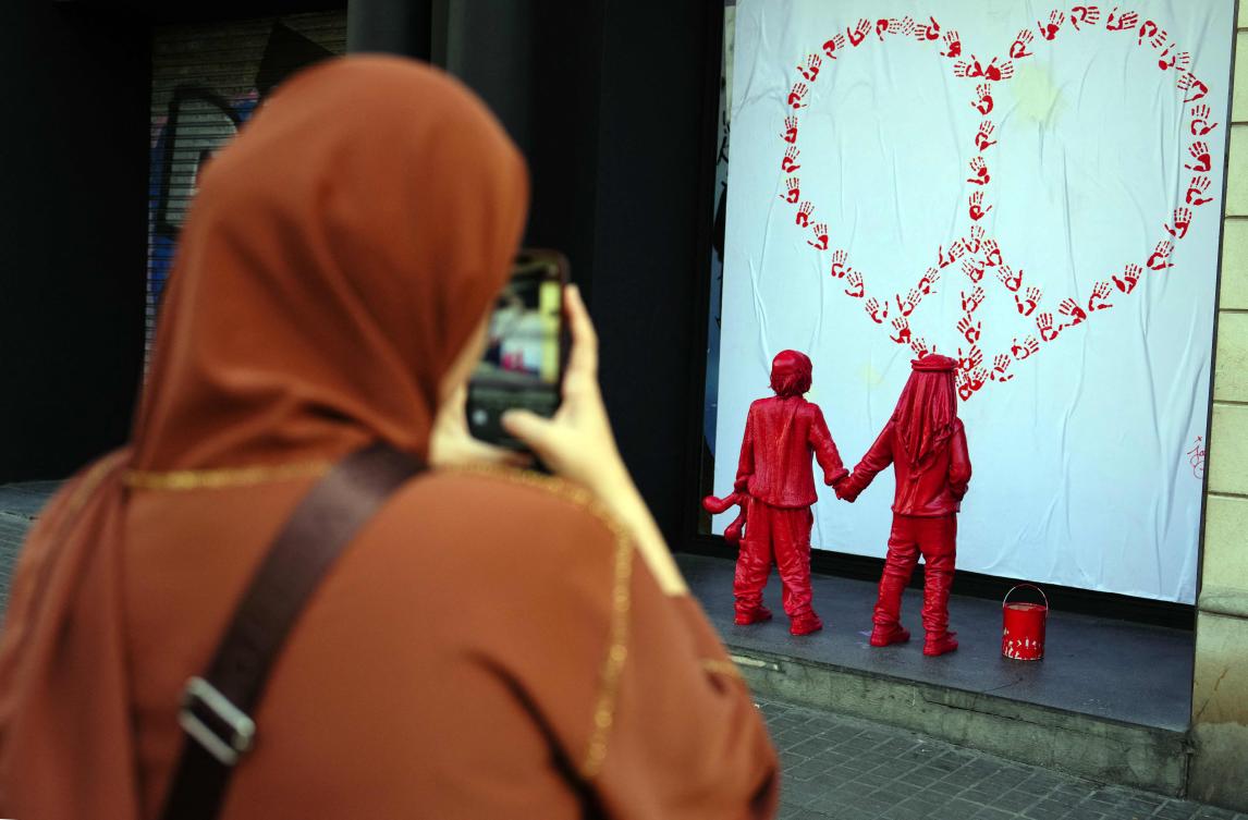 A woman takes images of a newly installed sculpture depicting two kids holding hands, one wearing a Jewish kippah and the other wearing a Palestinian keffiyeh, looking at a heart-shaped CND peace sign made with red hand prints, in Barcelona on Feb. 8, 2024. (AFP)