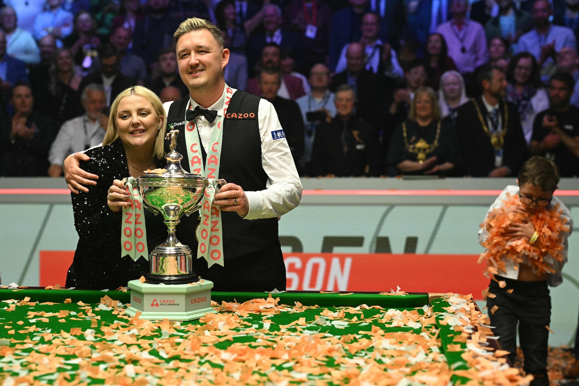 England's Kyren Wilson poses with the trophy and his wife Sofie as his son Finley (R) collects the confetti after victory over Wales' Jak Jones on day two of their World Championship Snooker final at The Crucible in Sheffield, northern England on May 6, 2024. (AFP)