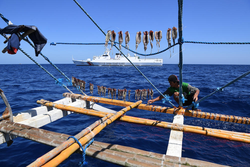 A Filipino fisherman dries squid on their fishing boat, while a Chinese coast guard ship monitors near the China-controlled Scarborough Shoal, in disputed waters of the South China Sea on Feb. 15, 2024. (AFP)