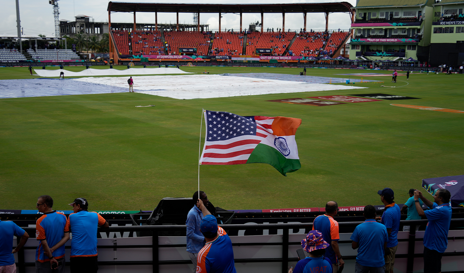 A fan waves a flag as the pitch area is seen covered after wet outfield delayed the toss for the ICC Men's T20 World Cup second semifinal cricket match between England and India at the Guyana National Stadium in Providence, Guyana, on June 27, 2024. (AP)