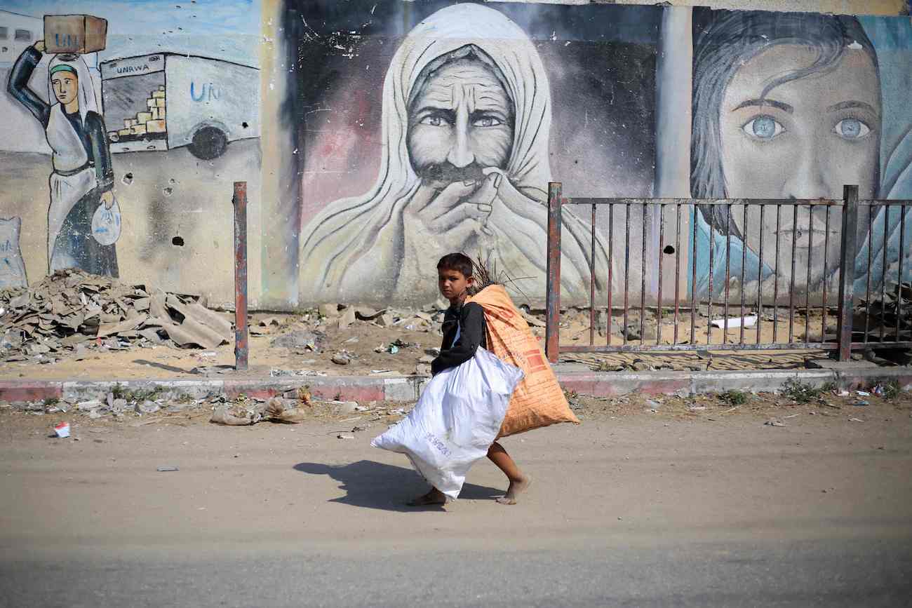 A displaced Palestinian child carries bags as he walks past mural paintings in Deir El-Balah in the central Gaza Strip on July 7, 2024. (AFP)