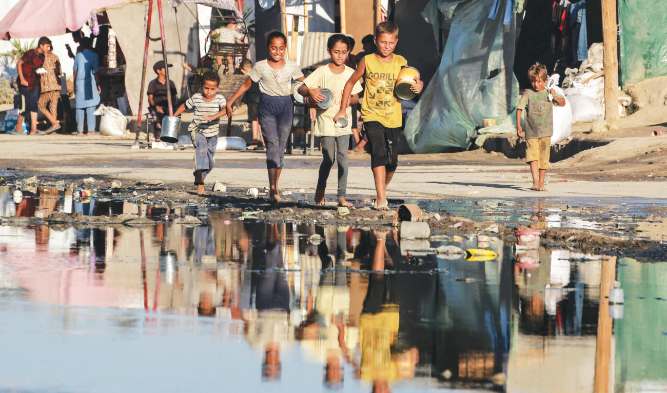 Palestinian children, carrying empty containers walk near stagnant wastewater, on their way to a food distribution point in Deir Al-Balah in the central Gaza Strip. (File/AFP)
