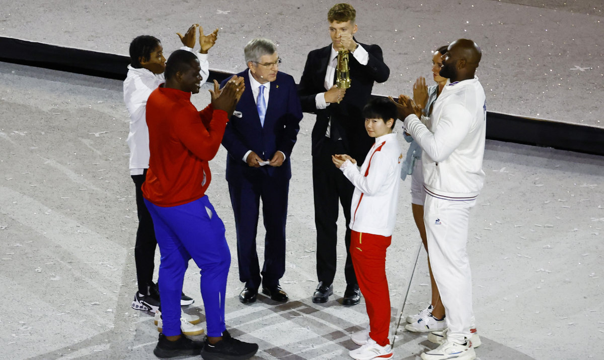 French swimmer Leon Marchand holds the Olympic flame lantern next to IOC President Thomas Bach, with athletes Teddy Riner of France, Mijain Lopez Nunez of Cuba, Yingsha Sun of China, Djankeu Ngamba of Refugee Olympic Team, Eliud Kipchoge of Kenya and Emma McKeon of Australia on stage. (Reuters)