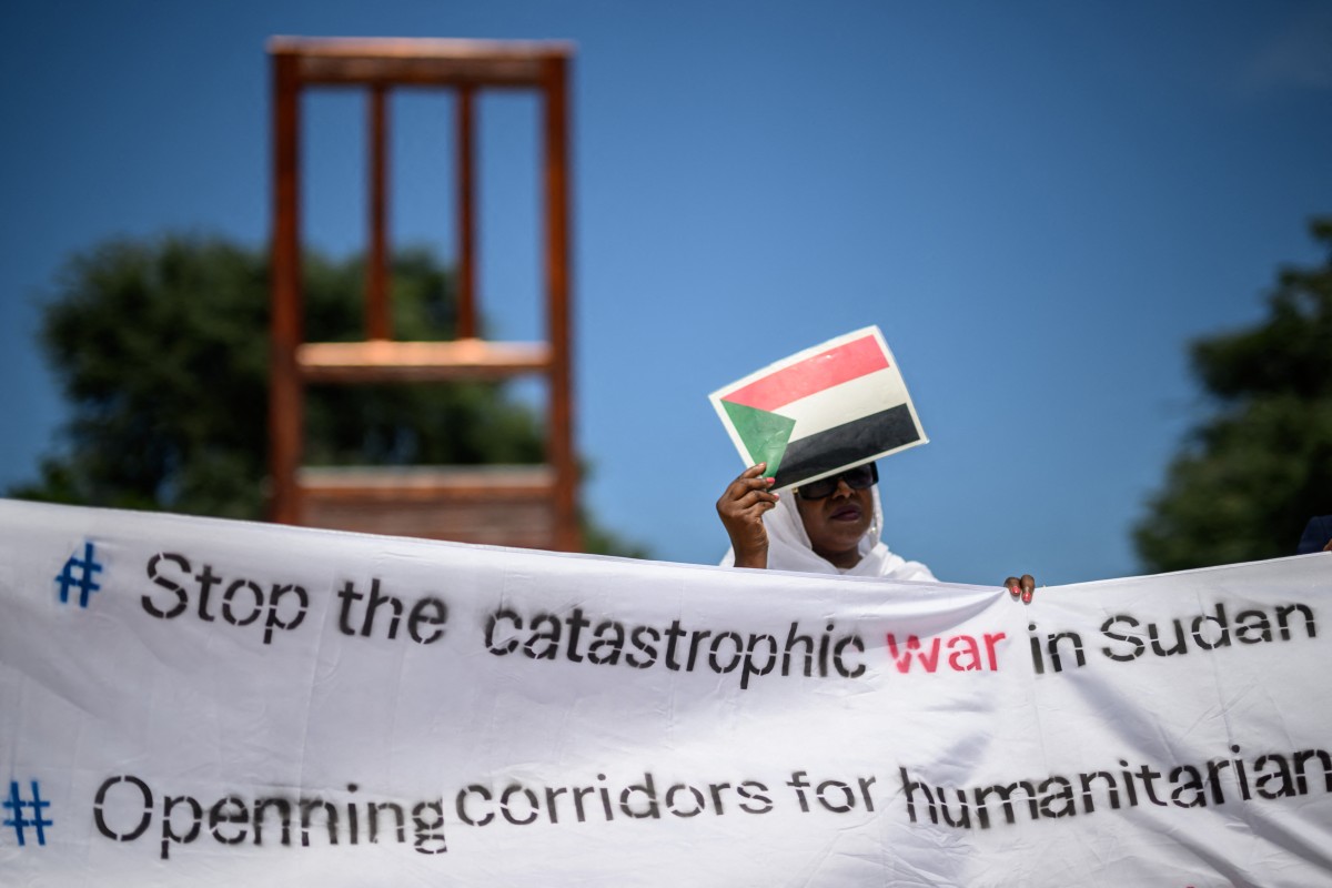 A woman protecting her face with a cardboard bearing the Sudanese flag as she holds a banner next to the monumental wood sculpture "Broken Chair" (L) during a demonstration on the opening day of Sudan ceasefire talks, in Geneva, on August 14, 2024 (AFP)