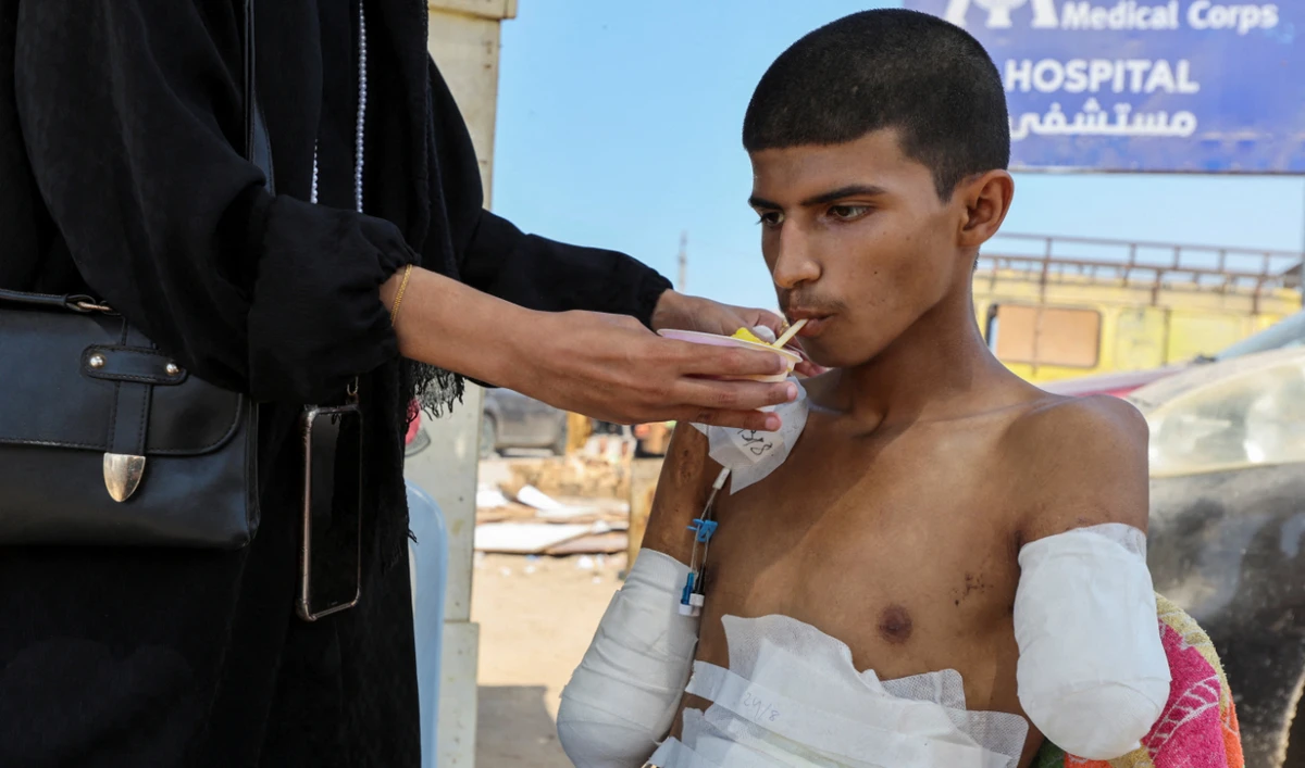 Palestinian teenager Diaa Al-Adini, who had his both arms amputated after being wounded in an Israeli strike on August 13 and was transferred from Al-Aqsa hospital due to an Israeli evacuation oder, is helped by his sister Aya to drink iced juice on a beach outside a field hospital, in Deir Al-Balah in the central Gaza Strip, August 27, 2024. (Reuters)