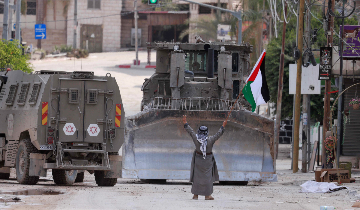 A Palestinian man lifts a national flag and flashes the victory sign as Israeli armoured vehicles including a bulldozer drive on a street during a raid in Tulkarem on September 3, 2024, amid a large-scale military offensive launched a week earlier in the occupied West Bank. (AFP)