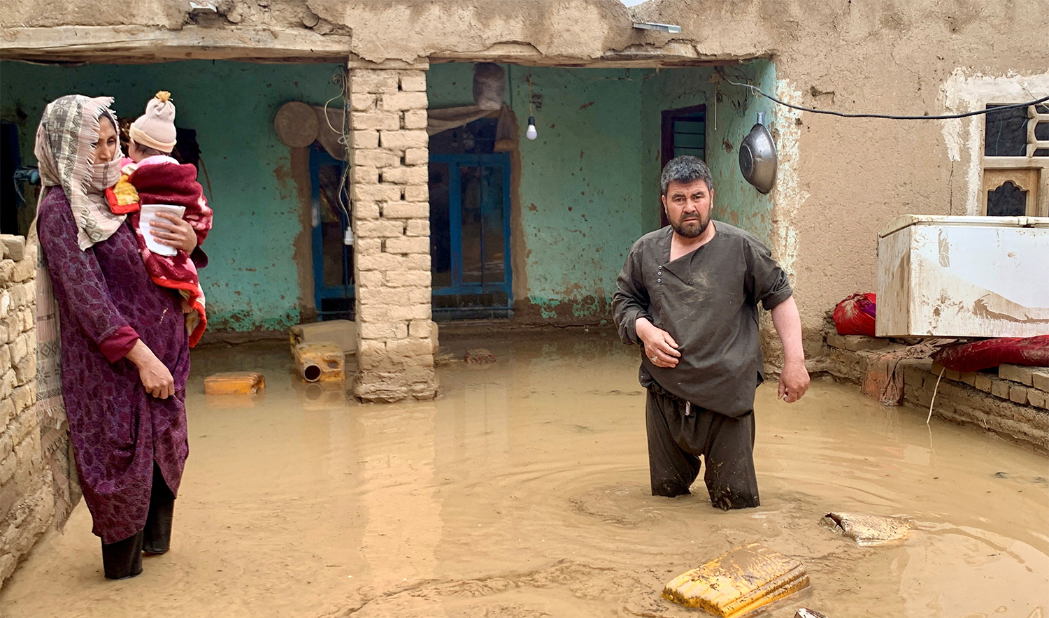 An Afghan woman holds her child as a man salvages his belongings from flood waters outside their house in Maymana, the capital city of Faryab Province, Afghanistan, on May 19, 2024. (REUTERS)