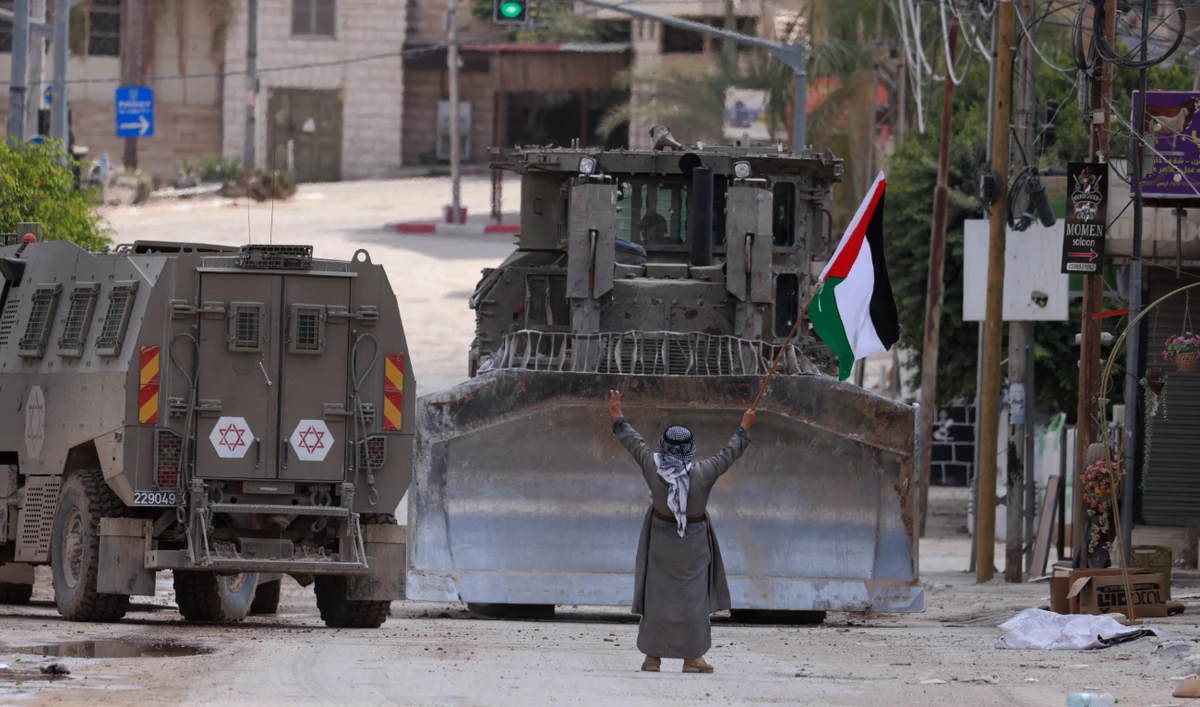 A Palestinian activist lifts a national flag and flashes the victory sign as Israeli armoured vehicles including a bulldozer drive on a street during a raid in Tulkarem on September 3, 2024. (AFP)
