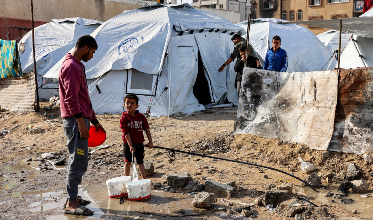 A youth speaks to a boy filling up buckets with water from a hose near tents at a shelter for displaced Palestinians in Gaza City on December 11, 2024. (AFP/File)